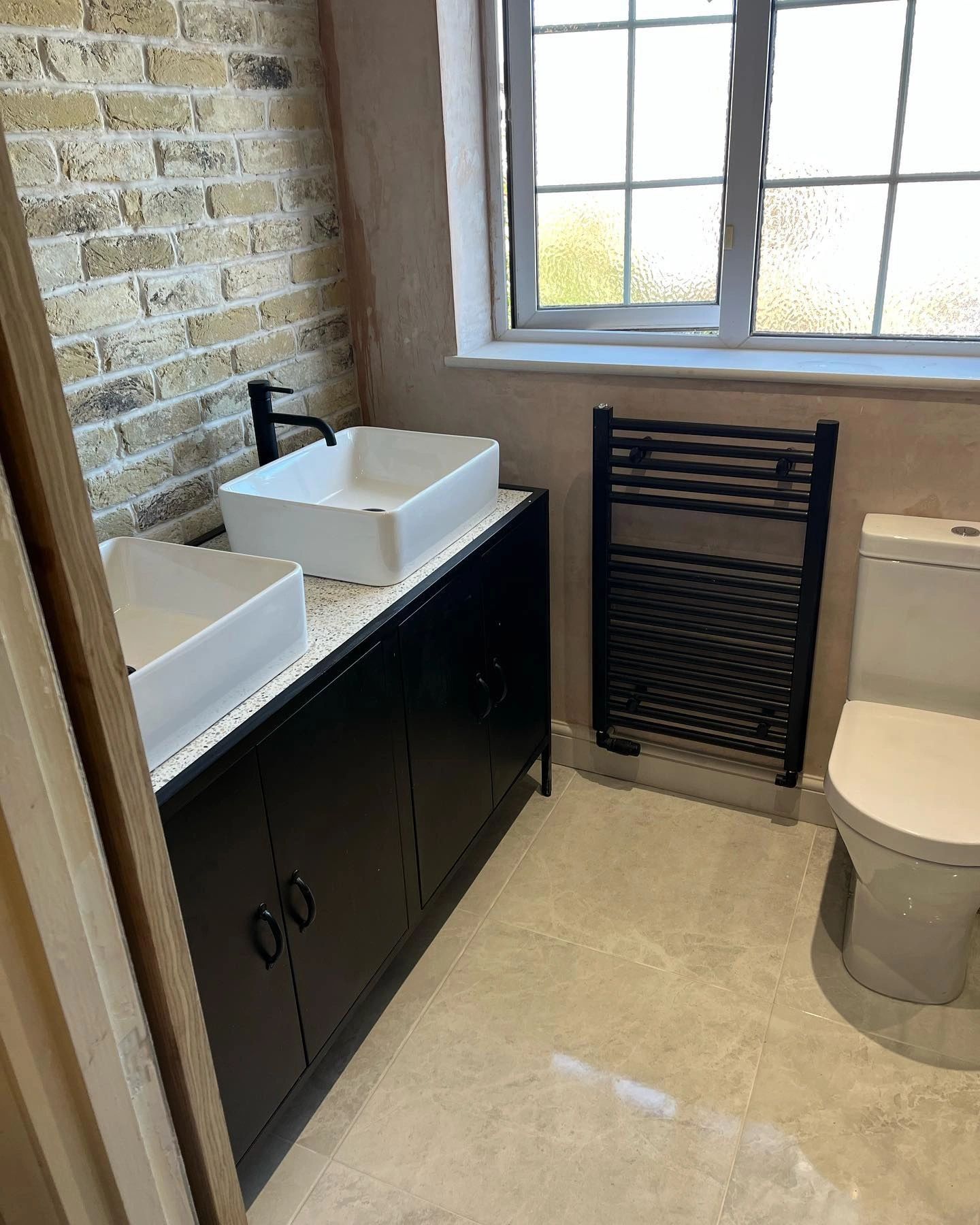 Fitted bathroom with twin countertop basins, black vanity unit, matt black towel radiator and exposed brick wall, installed by Acorn